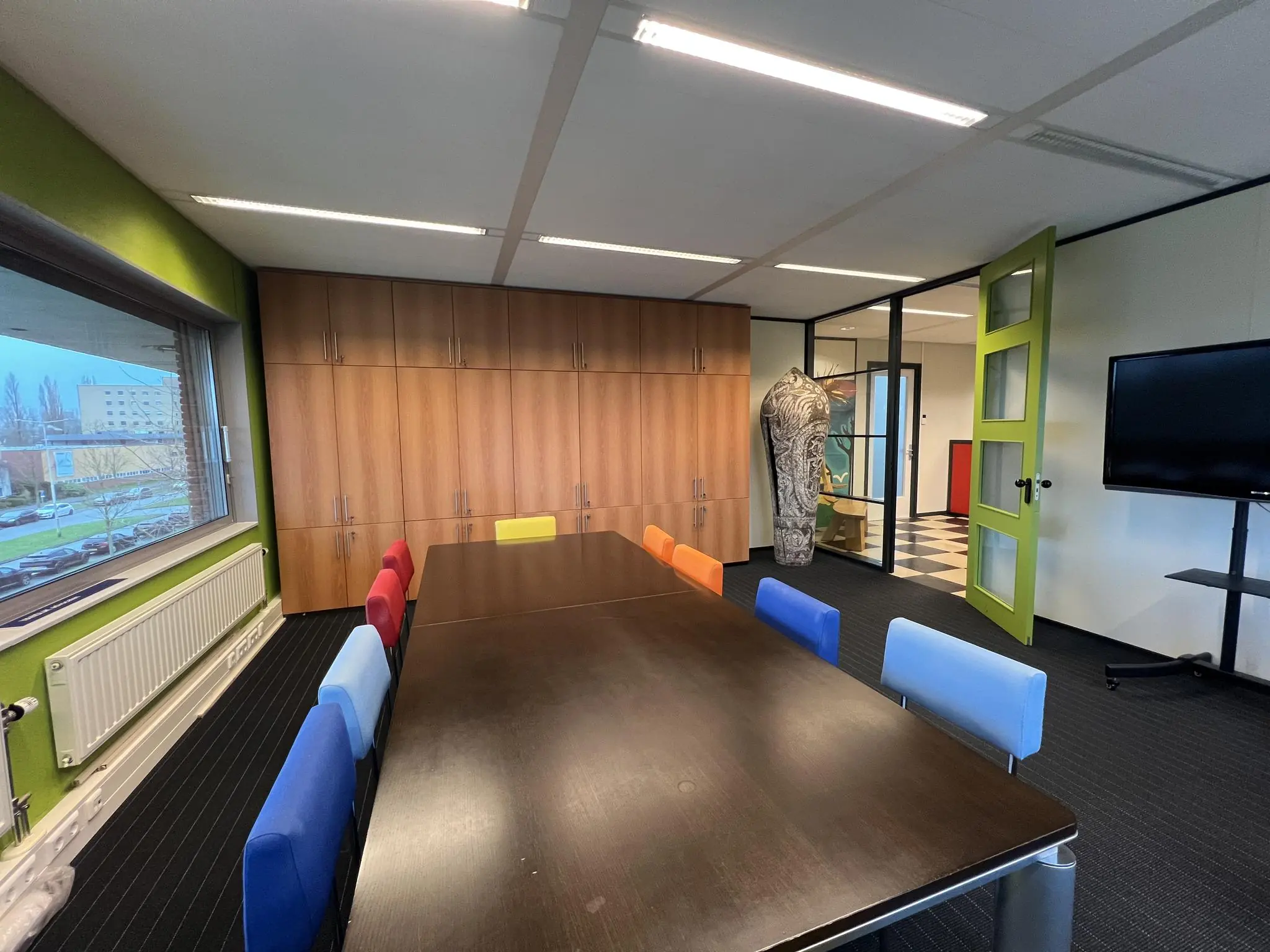 Vergaderruimte aan de Jacques Dutilhweg met kleurrijke stoelen, een grote tafel, houten kastenwand en uitzicht op de straat.